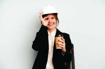 Young architect English woman with helmet and holding blueprints isolated on blue background excited keeping ok gesture on eye.