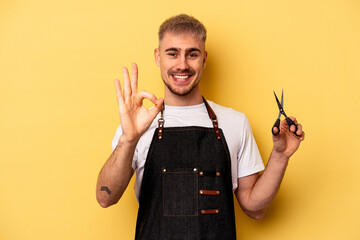 Young caucasian hairdresser man holding scissors isolated on yellow background cheerful and...