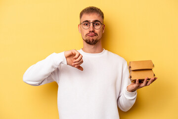 Young caucasian man holding a burger isolated on yellow background showing a dislike gesture,...