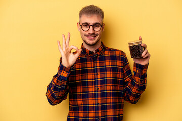 Young caucasian man holding coffee jar isolated on yellow background cheerful and confident showing ok gesture.