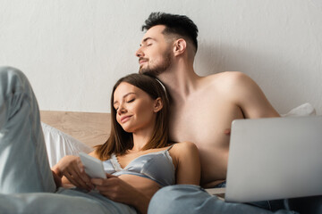 shirtless man and sexy woman in bra and jeans holding gadgets while relaxing with closed eyes in bedroom.