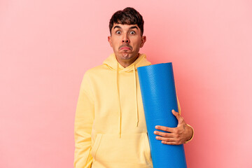 Young mixed race man holding a sport mat isolated on pink background shrugs shoulders and open eyes...