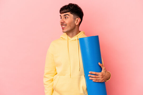 Young mixed race man holding a sport mat isolated on pink background looks aside smiling, cheerful and pleasant.