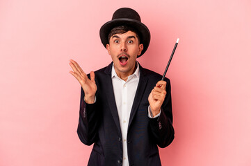 Young caucasian wizard man holding a wand isolated on pink background surprised and shocked.