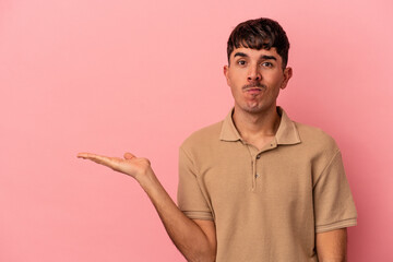 Young mixed race man isolated on pink background showing a copy space on a palm and holding another hand on waist.