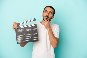 Young caucasian actor man holding clapperboard isolated on blue background looking sideways with...