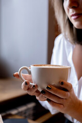 Young woman in white shirt sits in cafe and holds cup of coffee in hands. Lunch break