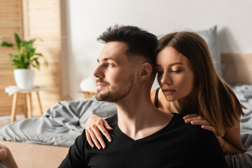 Obraz premium sensual young woman hugging shoulders of man in black t-shirt looking away in bedroom.
