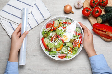 Woman putting plastic food wrap over bowl of fresh salad at wooden table, top view
