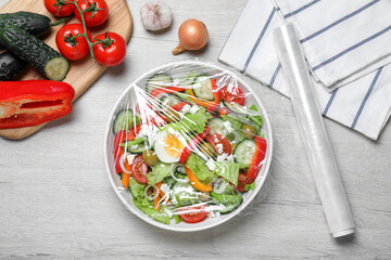 Bowl of fresh salad with plastic food wrap on white wooden table, flat lay - obrazy, fototapety, plakaty