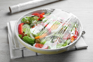 Bowl of fresh salad with plastic food wrap on white wooden table, closeup