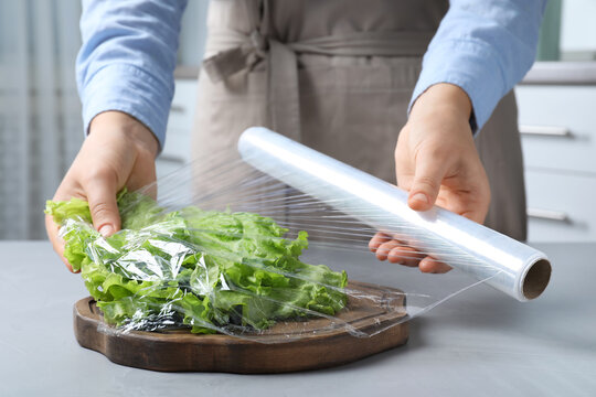 Woman putting plastic food wrap over fresh lettuce at light grey table indoors, closeup