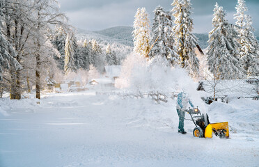 Unrecognizable person clearing off snow with snowblower in mountain village in a sunny winter day