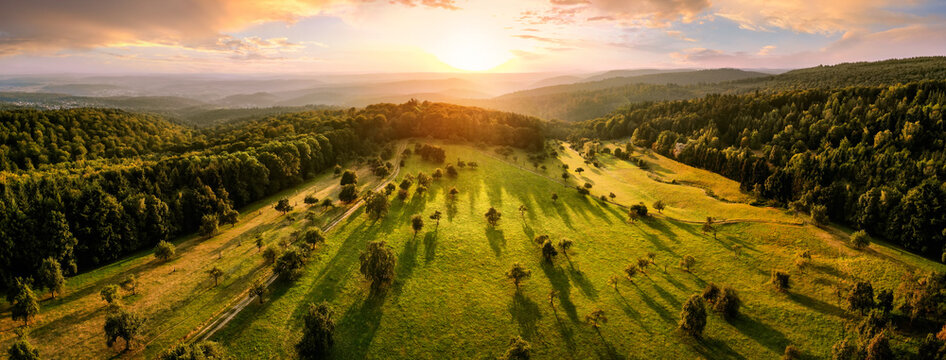 Aerial landscape panorama after sunrise: gorgeous scenery with the sun, trees on meadows in warm light casting long shadows, surrounded by forests
