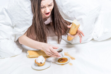 The girl is indignant, spilled coffee on the bed. Breakfast. The brunette lies in bed, next to her is a cupcake, a baguette in her hand and a cup