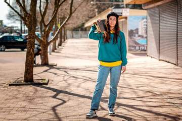 A young Caucasian hipster woman poses with a skateboard over her shoulders. In the background, an alley. Concept of sports lifestyle and street culture