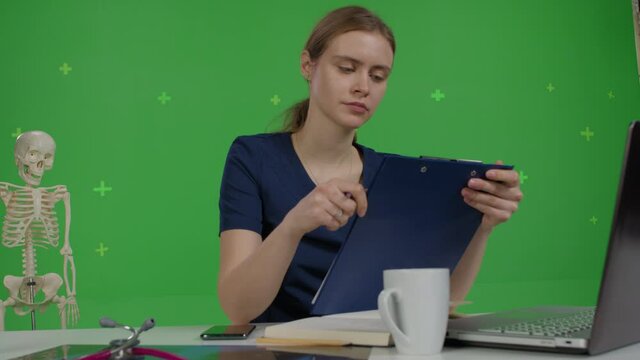 Caucasian female doctor sits at her desk, looks into a laptop, talks with a patient. A medical student listens to a lecture or communicates with teacher over green screen background. Perspective view