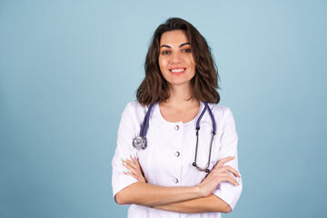 Young beautiful woman doctor in a lab coat on a blue background looks at the camera with her arms crossed and smiles confidently