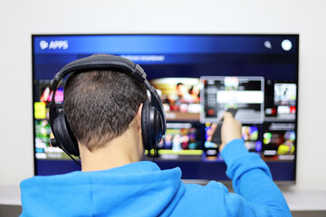 Man in headphones sitting with remote controller on smart TV screen background. Person choosing...