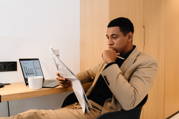 Black man reading newspaper while working with laptop at desk