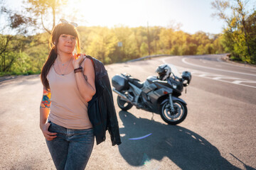 Fototapeta na wymiar Stylish tattoed girl with a leather jacket poses against the background of a motorcycle by the road. Motorcycle local travel concept and World Motorcyclist Day