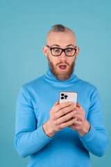 A young man with a red beard in a turtleneck on a blue background holds a phone and looks at the screen in shock and surprise