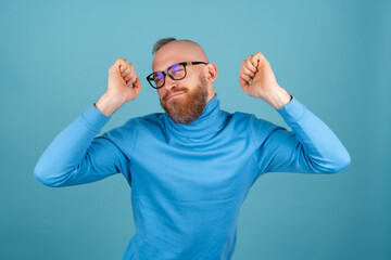 A young man with a red beard in a turtleneck on a blue background is dancing merrily, having fun