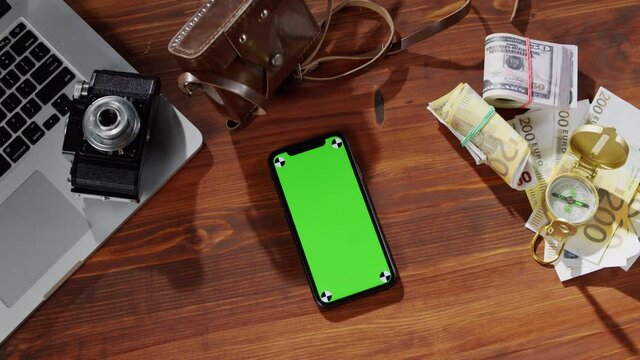 Smartphone with chroma key top view, money cash, compass, photo camera and laptop computer on table. Traveling and tourism, mobile phone with green screen close-up.