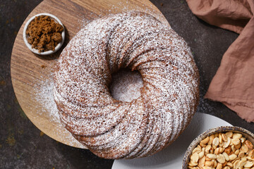 Muffin or bundt cake decorated icing sugar on the stand and spoon with brown cane sugar and peanuts in a bowl on the gray table. Shallow depth of field