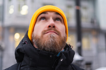 A young man with a red beard in an orange hat in the winter on a proud street in the evening looks up into the sky