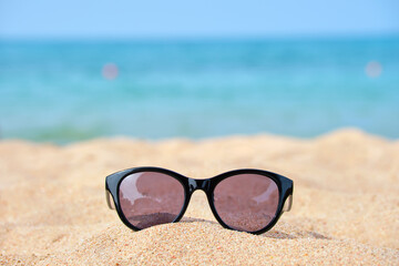 Fototapeta na wymiar Closeup of black protective sunglasses on sandy beach at tropical seaside on warm sunny day. Summer vacation concept
