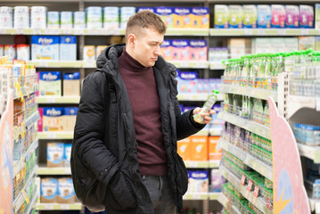 Photo of a handsome brunet young man in a burgundy sweater who chooses baby food in a store in a mall. Shopping concept