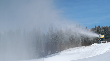 Obraz na płótnie Canvas Snow cannon blows artificial snow on mountain slope, background of forest, blue sky in ski resort on sunny day. Snowgun makes snow of water in cold winter. Snowmaker sprays water on mountainside