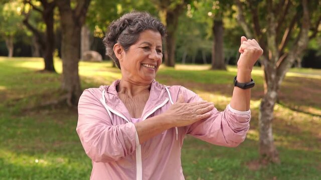 Happy cheerful Elderly brown woman with pink jacket flexing arms, showing strength and joy. Outdoor in green area. Active, wellness, leisure, freshness concept
