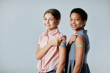 Portrait of two cute girls showing shoulder patch and smiling at camera after getting vaccinated,...