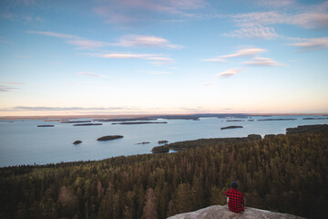 View of an adventurer in red and black shirt sitting on the edge of a cliff watching Lake Pielinen...