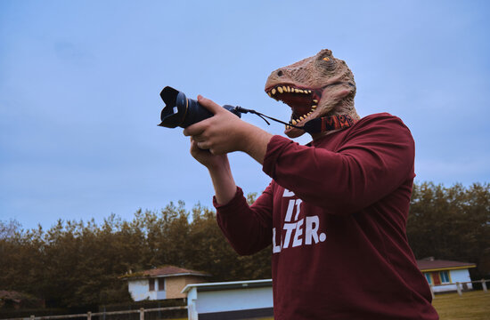 Hombre disfrazado con una máscara de dinosaurio en un parque con una cámara de fotos en la mano