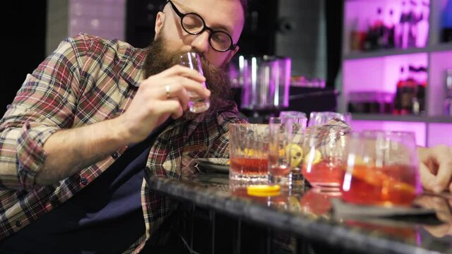 A very drunk young man is lying with his head on a table in a bar. A drunk man in depression sleeps, wakes up and drinks a glass. There are a lot of empty glasses on the table next to the client.