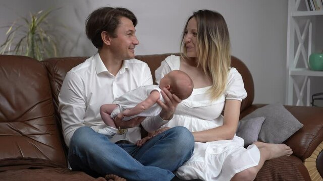 Portrait of happy couple sitting on couch with newborn baby admiring child and looking at camera smiling. Wide shot positive Caucasian husband and wife posing at home with infant son in slow motion