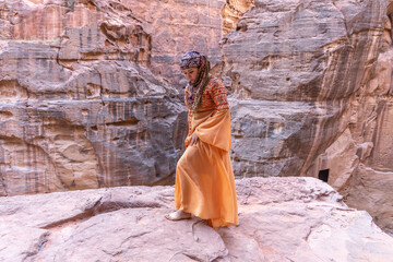 woman dressed in traditional clothes and headscarf staying at the top of rock on the background is Petra palace Jordan - obrazy, fototapety, plakaty