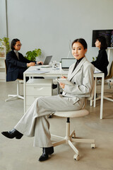 Minimal full length portrait of elegant Asian businesswoman looking at camera while sitting on chair in office
