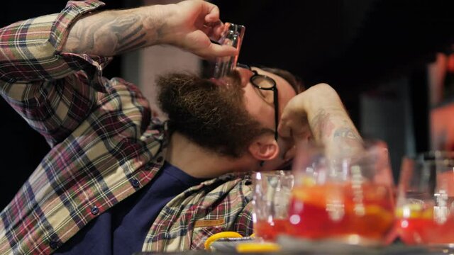 A very drunk young man, a man with glasses and a beard, is lying with his head on a table in a bar. A drunk man in depression sleeps, wakes up and drinks a glass. Lots of empty glasses.