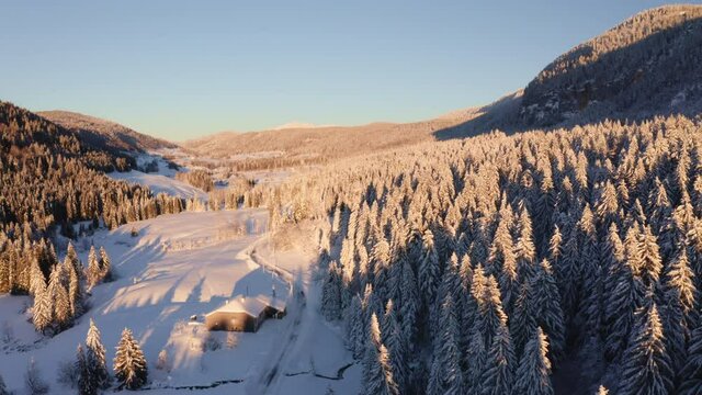 Survol drone au soleil couchant d’une vallée de montagne recouverte de neige. Lumière chaude rasante sur la foret de sapins du Jura avec route et maison recouverte de neige sur le toit, Valserine