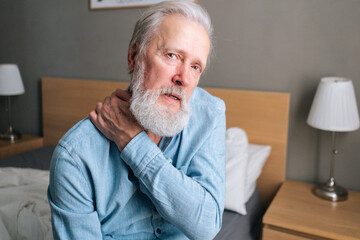 Close-up of senior adult man warming up and massaging shoulder sitting on bed in morning after waking up at home, looking at camera. Bearded mature man suffering from muscle tension after waking up.