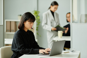 Side view portrait of young Asian woman using laptop at desk in office cubicle, copy space