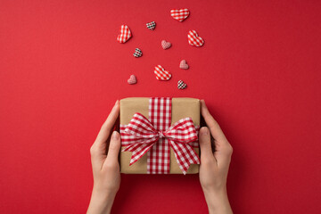 First person top view photo of valentine's day decorations girl's hands holding kraft paper giftbox with checkered ribbon bow and hearts on isolated red background