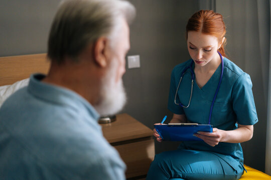 Close-up view from back of sick senior male patient talking with female doctor writing down diagnosis and symptoms in medical history on clipboard at home. Elderly man getting medical consultation.