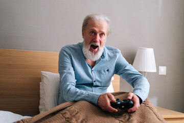 Portrait of screaming gray-haired senior male playing online video game using gamepad at home. Happy bearded mature elderly man in pajama playing computer game having fun with joysticks. © dikushin