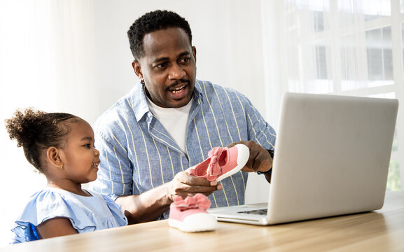 Happy black handsome man with his little daughter using laptop for shopping shoes and cloth online at home. Being a new baby in mixed race family. - Powered by Adobe