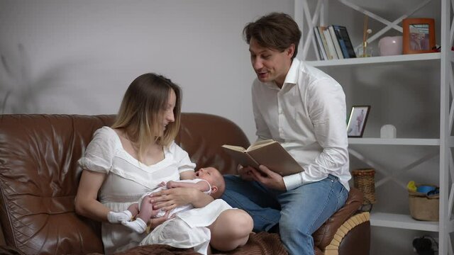 Portrait Of Smiling Father Singing Lullaby Reading Book Sitting With Wife And Newborn Son On Couch At Home. Cheerful Caucasian Man Enjoying Infant Nap Time With Woman And Baby Boy In Slow Motion